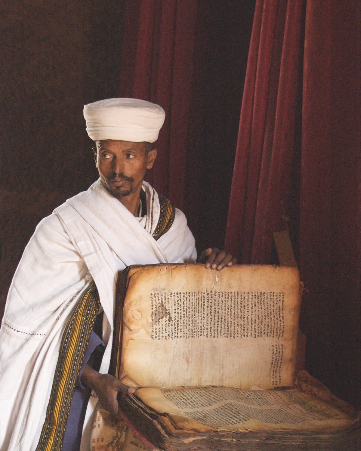 An Ethiopian Orthodox priest in white robes and turban holding an old manuscript open at Lalibela
