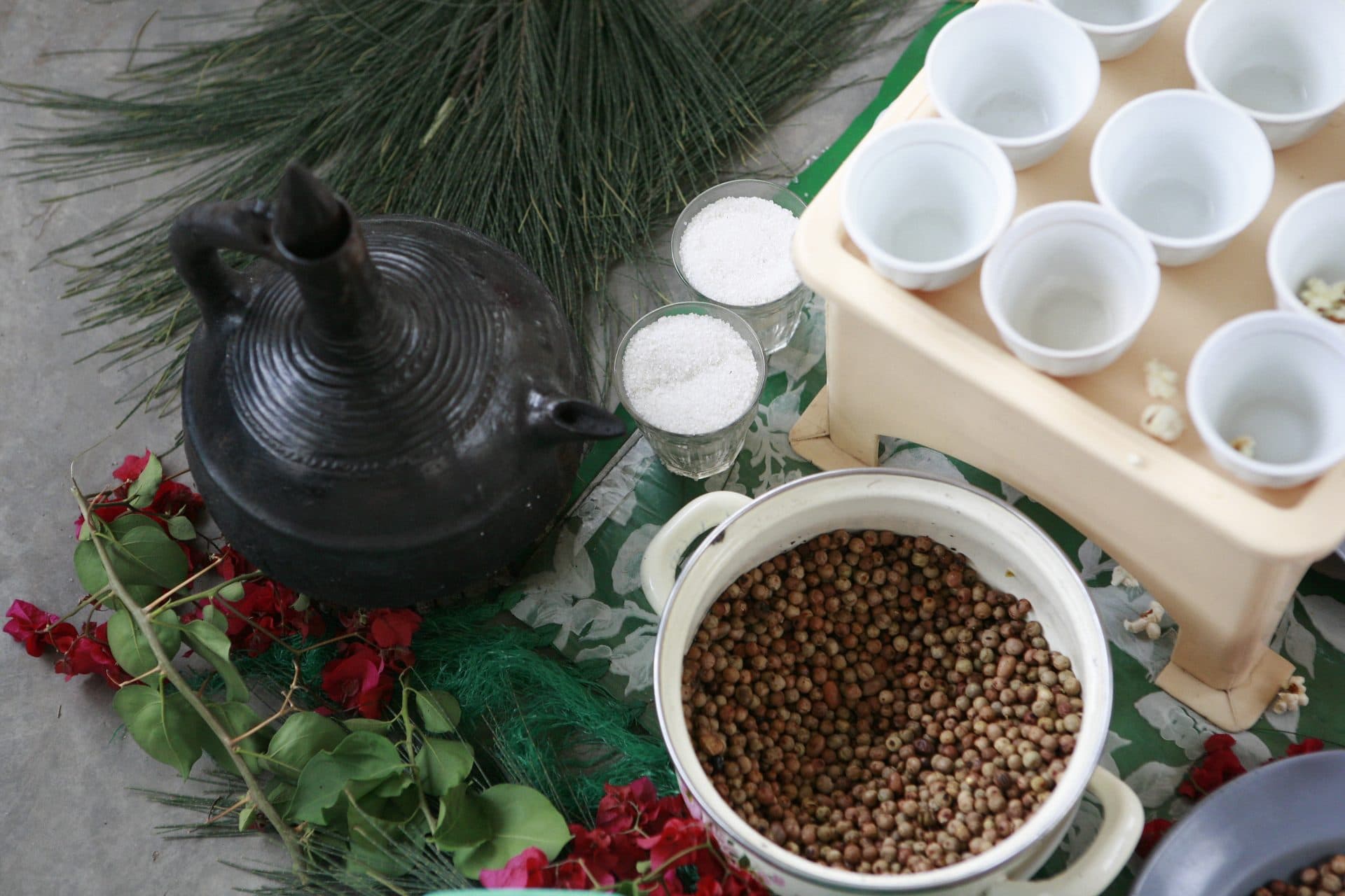 A black clay jebena beside a tray of small white cups and a bowl of roasted coffee beans, set on fresh grass and flowers