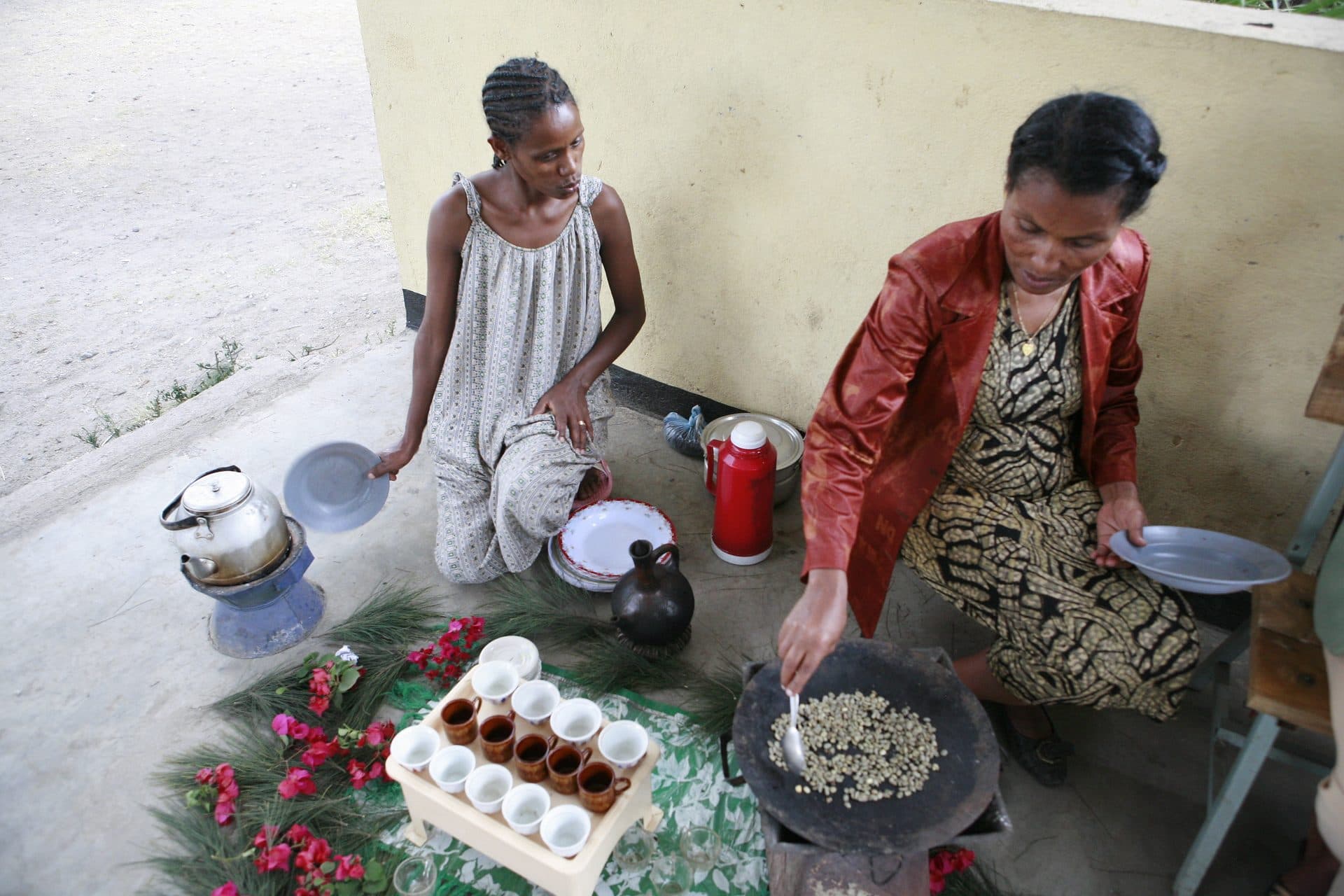 Two women seated on the floor roasting green coffee beans in a pan beside a tray of small cups and a jebena