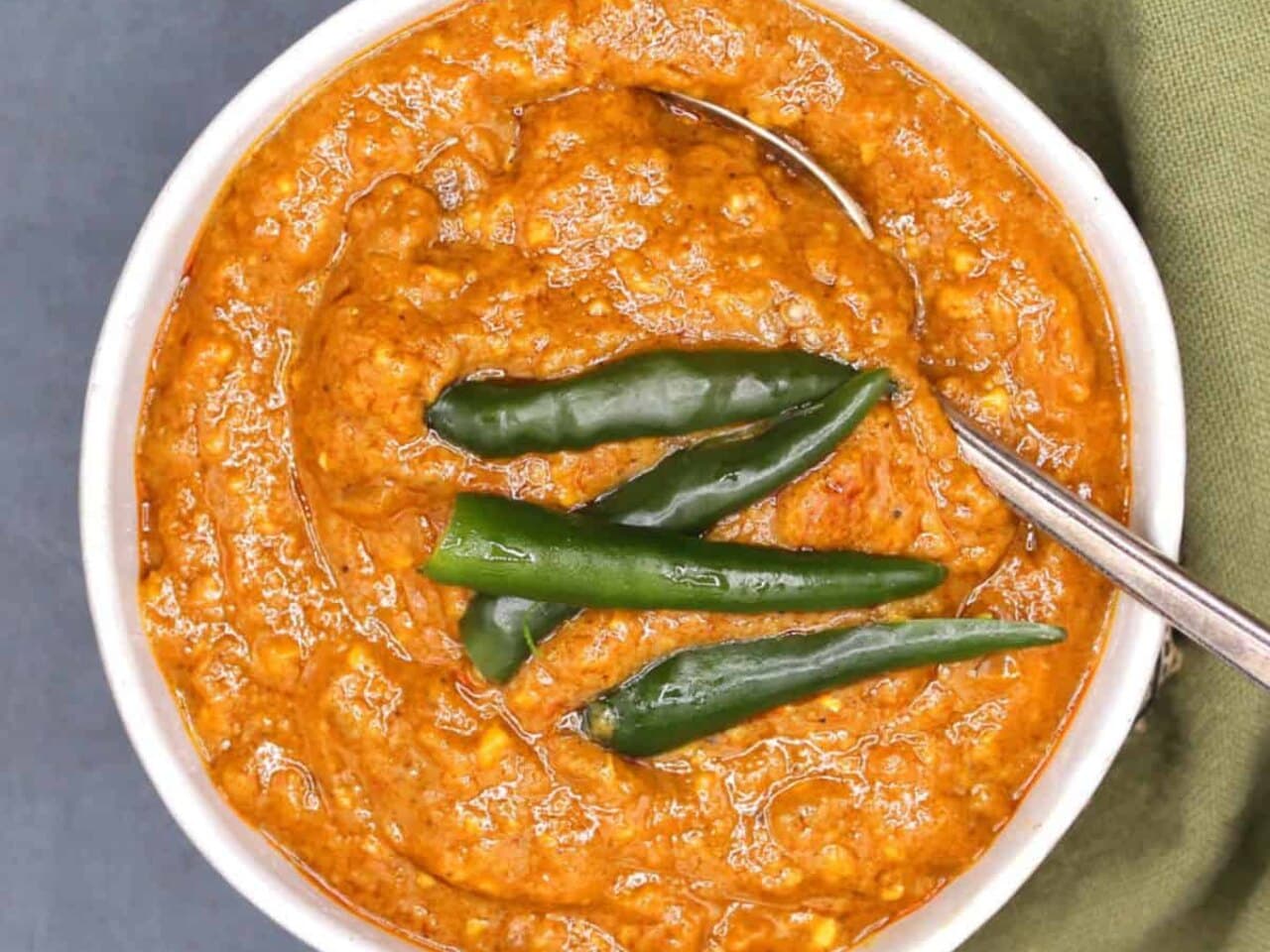 A white bowl of thick orange shiro topped with whole green chili peppers, a spoon resting in it