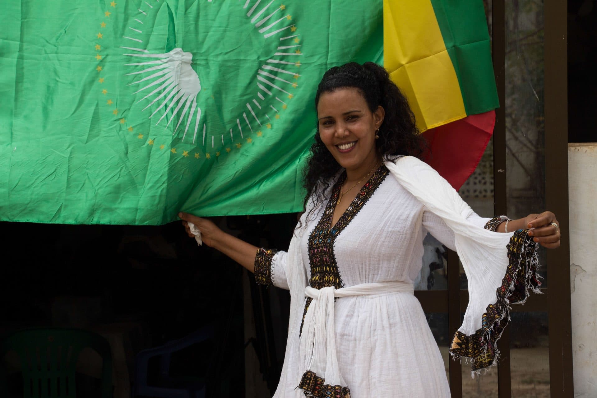 A smiling woman in a white habesha dress holding the flags of Ethiopia and the African Union