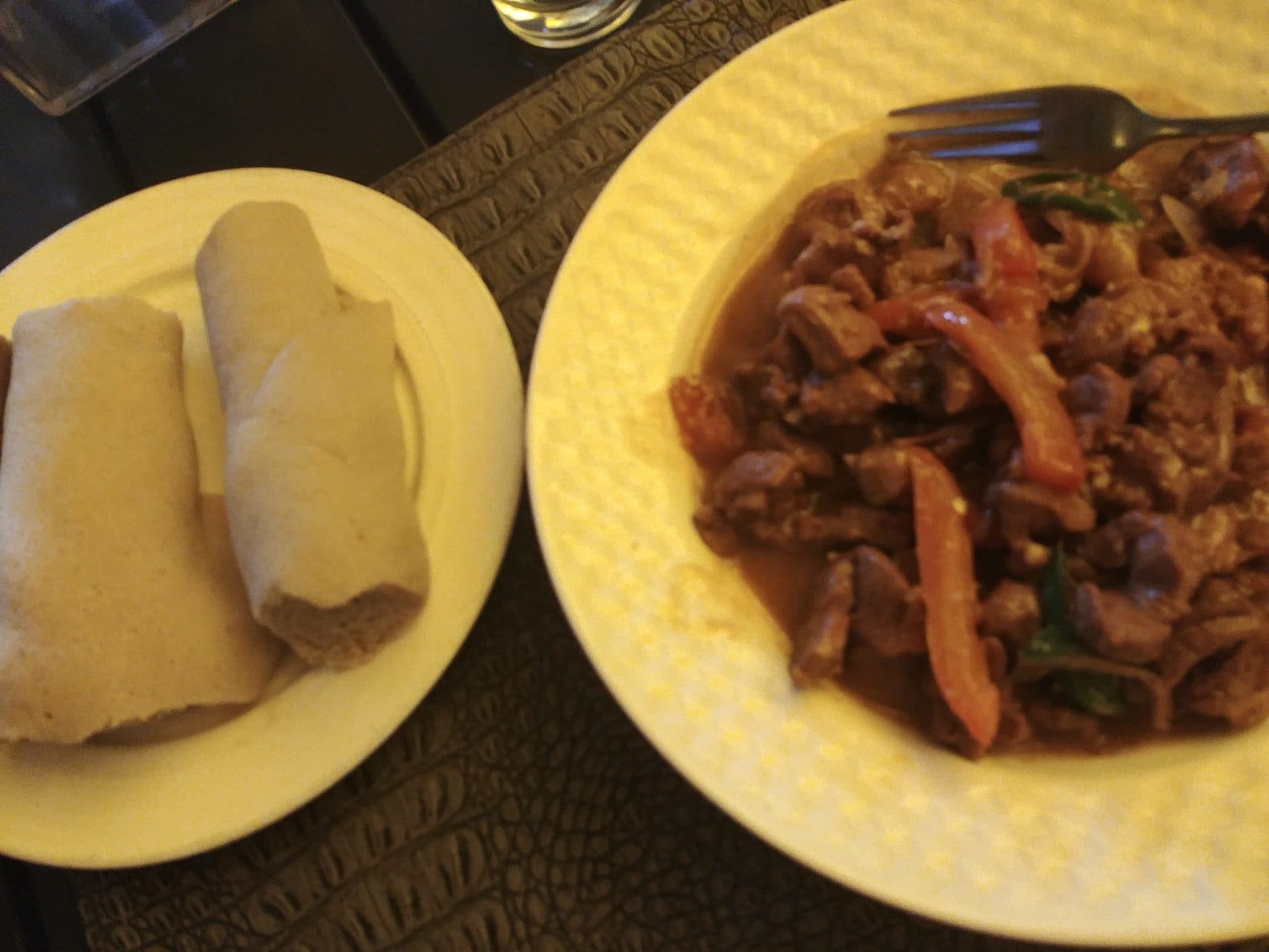 Two plates seen from above: rolled injera on one and mutton tibs with onions and peppers on the other