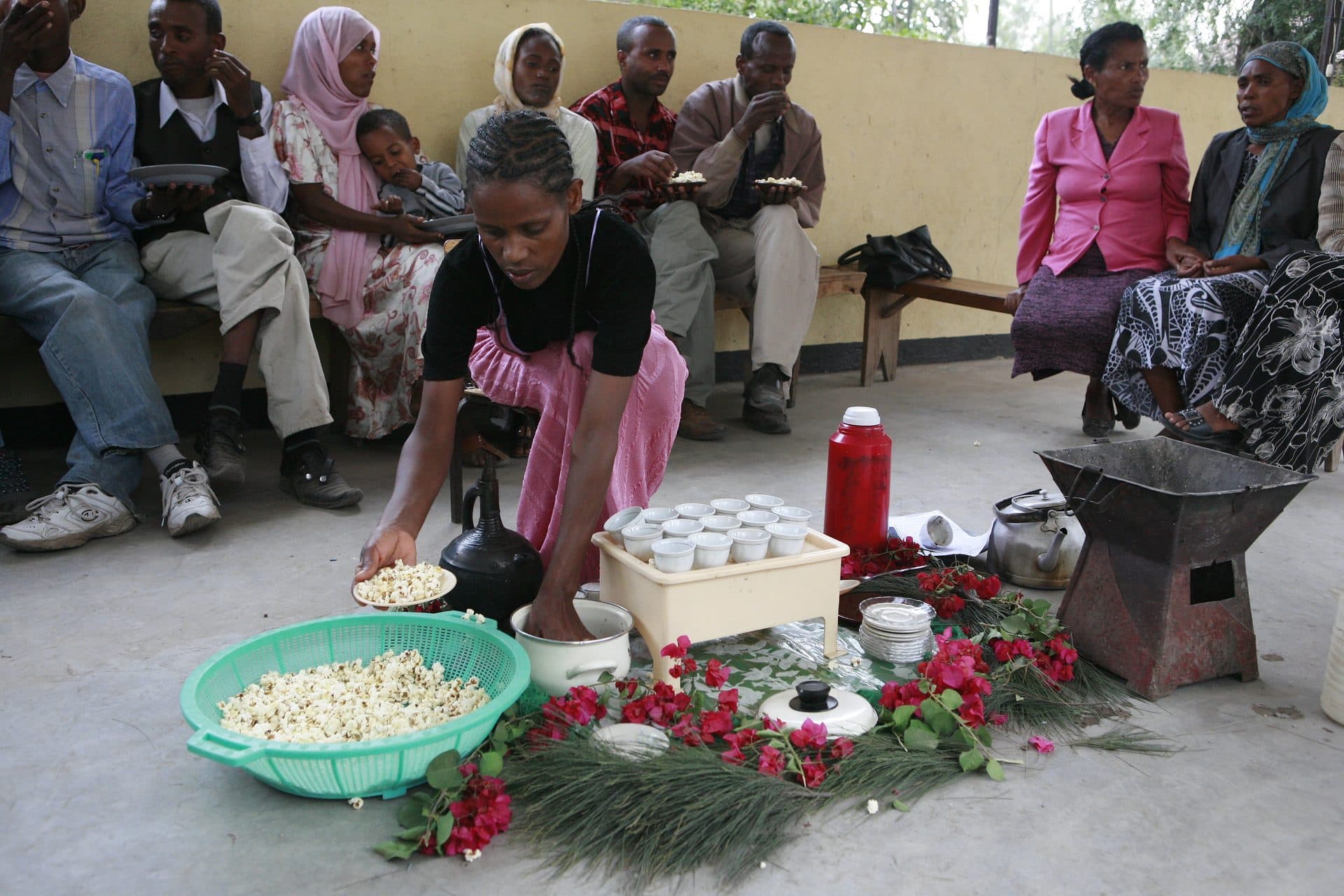 Neighbors seated along a wall watching as a woman prepares coffee on the floor, with cups, beans and fresh grass laid out for a coffee ceremony